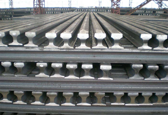 Multiple layers of heavy steel railway tracks stacked neatly in an industrial storage facility.