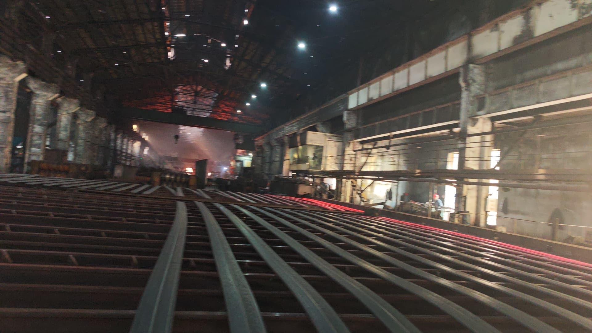 Rows of cooling steel rails in a dark industrial factory with glowing hot metal background.