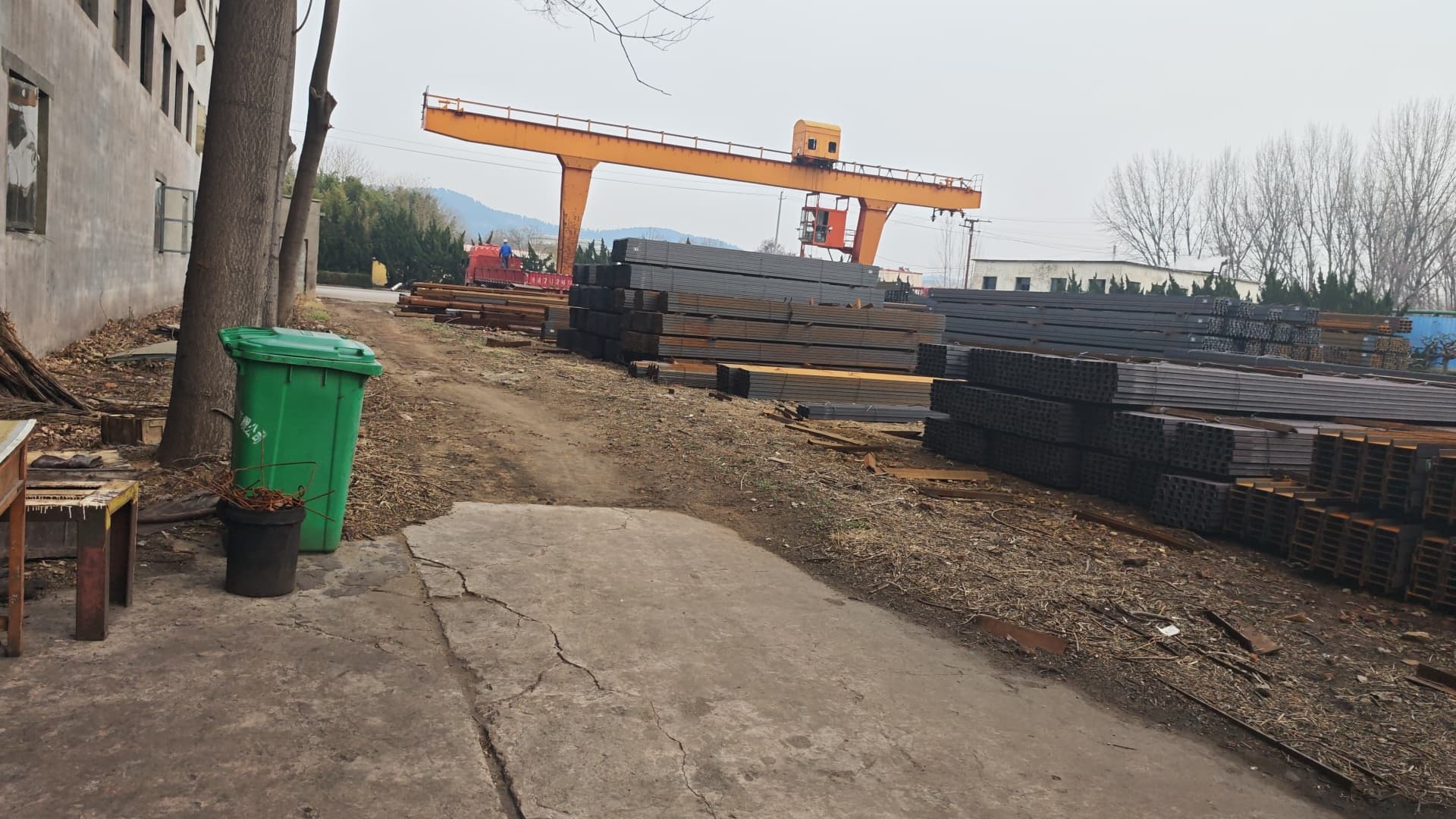 Industrial storage yard featuring an orange gantry crane and stacks of steel beams.