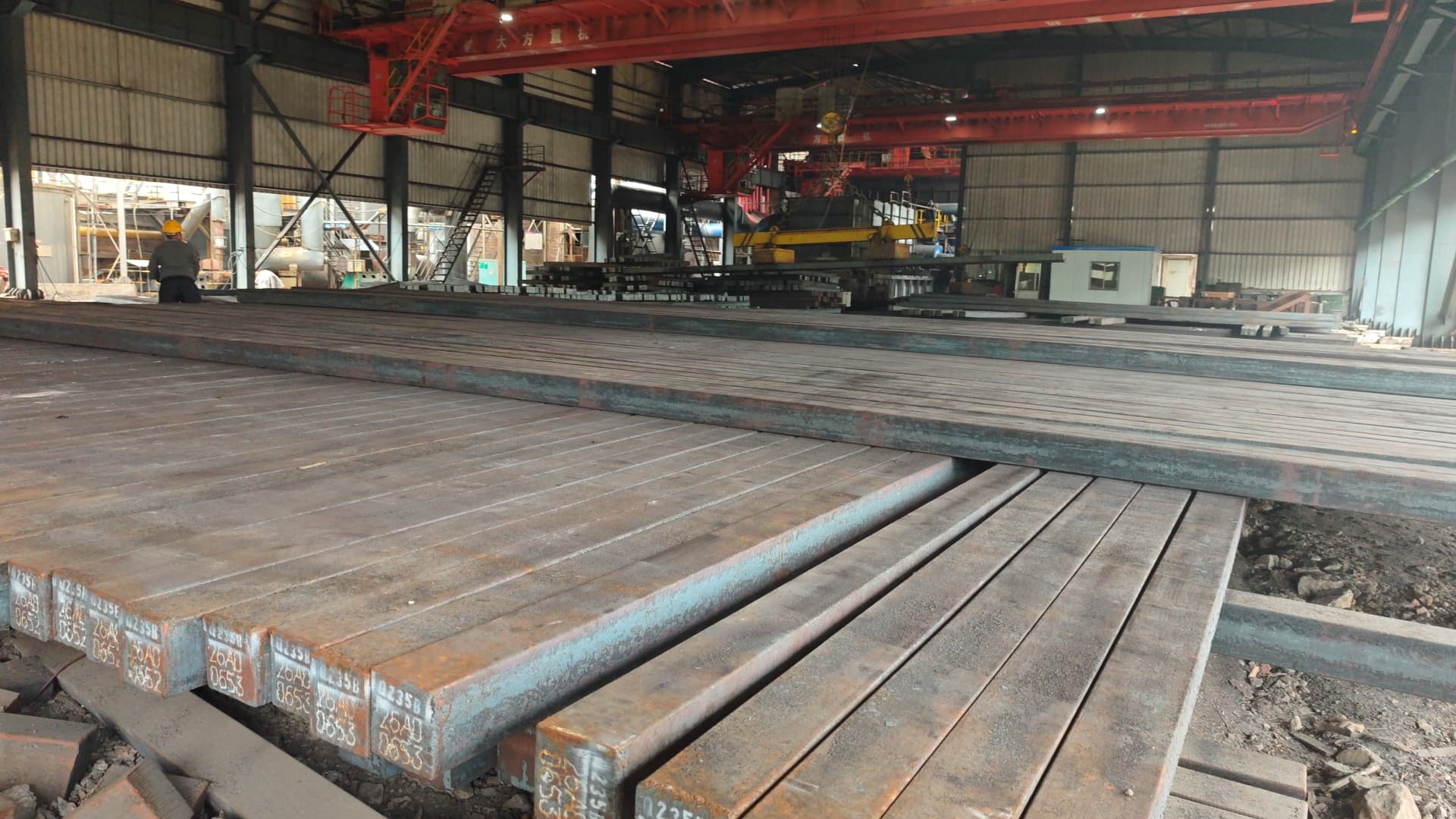 Stacked steel billets in an industrial warehouse with a red overhead crane and a worker.
